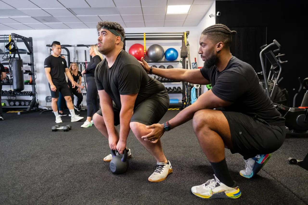 A Formula Fitness coach crouching beside a client who grips a kettlebell on the floor in a squat position, with other coaches and clients training in the background